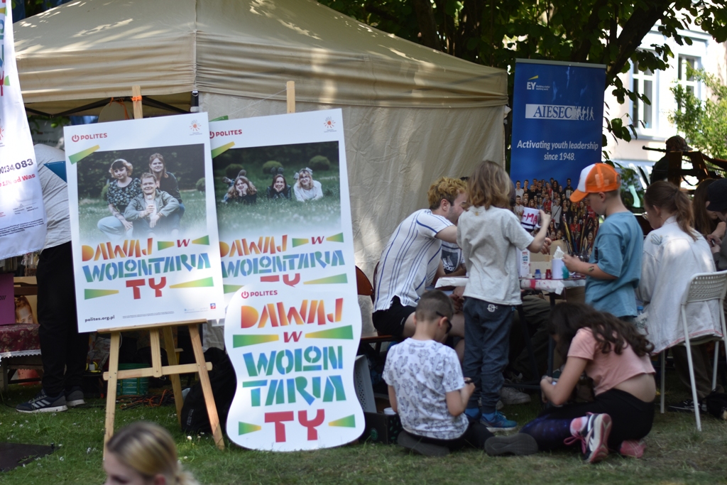 Zdjęcie z pikniku pasji - w tle namiot, przed namiotem sztalugi z plakatami. Obok stół przy którym warsztaty dla dzieci prowadzą przedstawiciele stowarzyszenia.