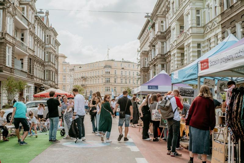 Zdjęcie ilustrujące wydarzenie. Na fotografii ludzie odwiedzający stoiska ustawione na ulicy Rayskiego w Szczecinie. Także dzieci bawiące się na rozwiniętej sztucznej trawie. Tłem fotografii są kamienice przy ul. Rayskiego. Autor zdjęcia - Adam Piaseczny. 