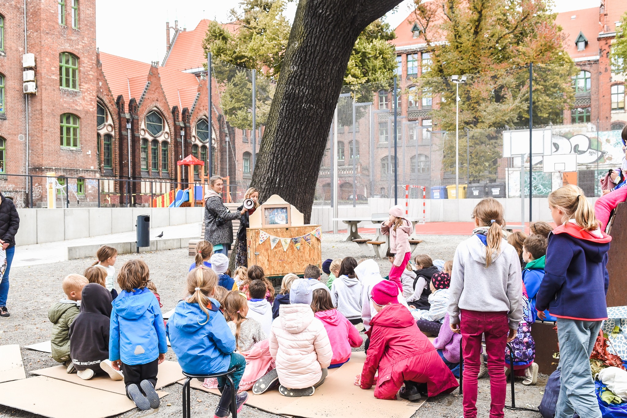 Zdjęcie organizatora autorstwa Moniki Lebery. Na boisku szkolnym przy SP 1 w Szczecinie grupa dzieci realizuje warsztaty. dzieci siedzą na kartonach. Pod drzewem stoją dwie osoby prowadzące warsztat. 